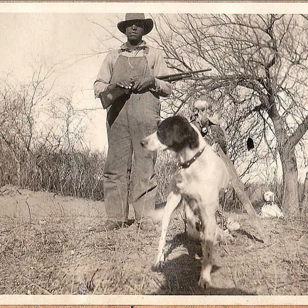 Early 1900s sepia print — a Black hunter in overalls and brimmed hat holding a shotgun, his setter in the foreground