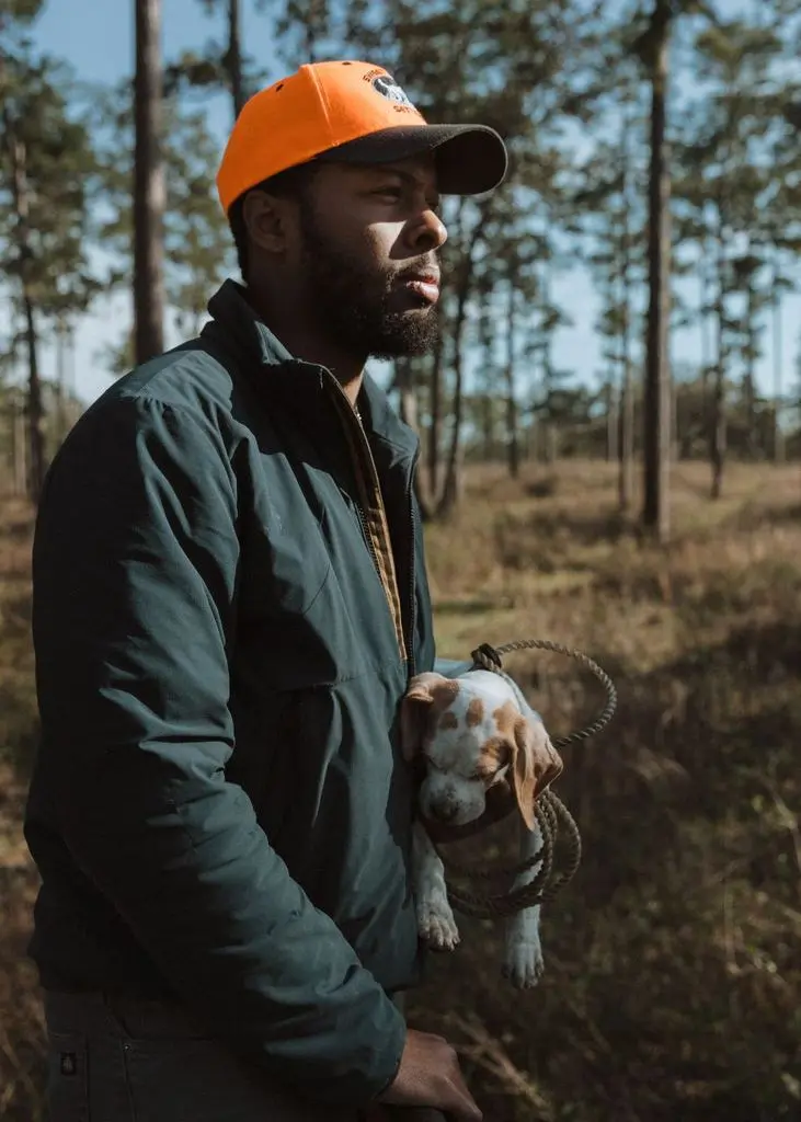 Handler in blaze orange carrying a pointer puppy through the longleaf
